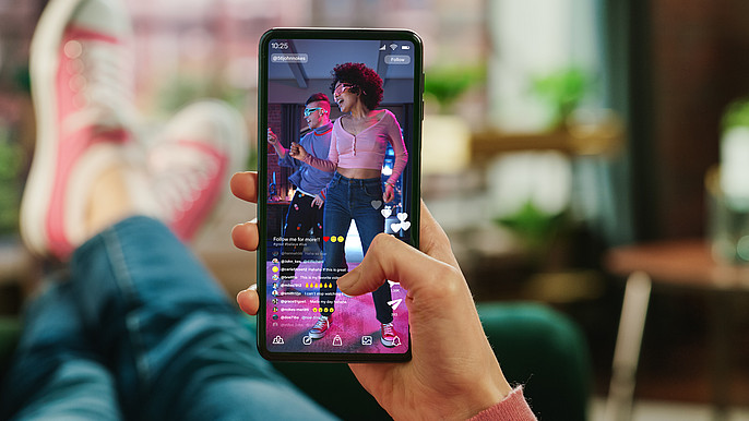 Eine weibliche Hand hält ein Handy auf dem eine Tanzszene zu sehen ist